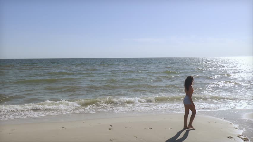 Beautiful young woman enjoying a peaceful barefoot walk along the beach under clear blue skies. Perfect for travel, wellness, or summer lifestyle stock video and creative promos.
