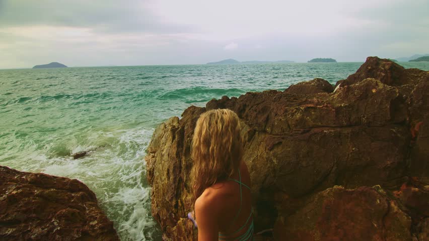 Sexy hot woman meditates, relaxes on rock crack reef hill in stormy morning cloudy sea. Girl in blue swimsuit. Concept feminine, sexual vaginal health, hygiene, womanly, freedom, freshness, recovery