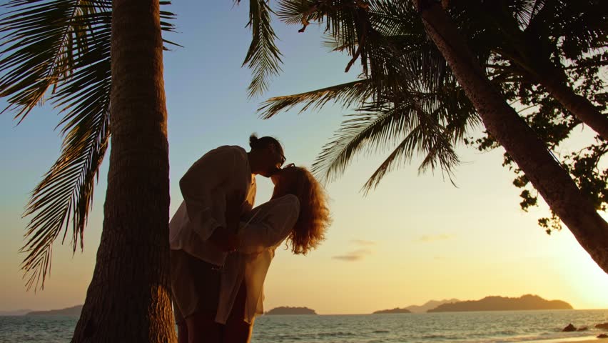 SILHOUETTE DARK GOLD SUNSET VIEW: Loving couple in white shirt and sunglasses, near palm tree, kissing, hugging, relax and drinking cocktail. Concept tropical summer vacations, holidays wedding love