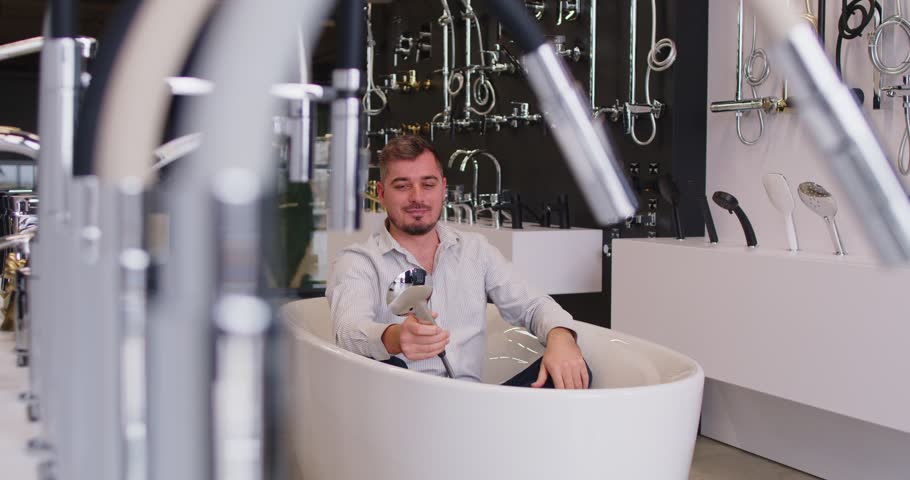 Young happy funny man customer choosing bathtub for bathroom in his house at building market or shopping mall planning repair. Man in plumbing shop having fun sitting in tub. 4k video.