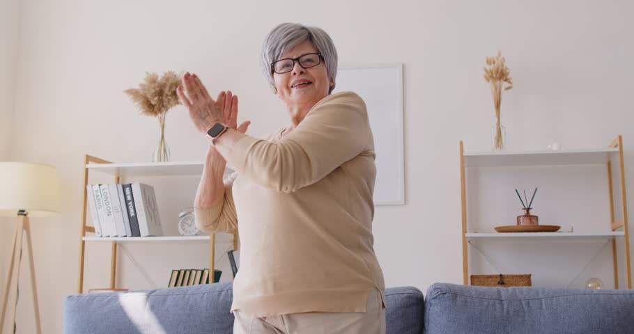 Cheerful energetic senior woman with gray hair dancing to music in living room and looking at camera. Happy smiling elderly lady enjoying movement freedom, feeling positive and full of vitality.