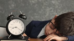 Cute school-age boy sleeping on desk with crooked glasses. Alarm clock shows 7 AM. Child in jacket resting head on notebook. Back to school concept, concrete wall background. - Powered by Shutterstock - Get 15% off with code: PIKWIZARD15