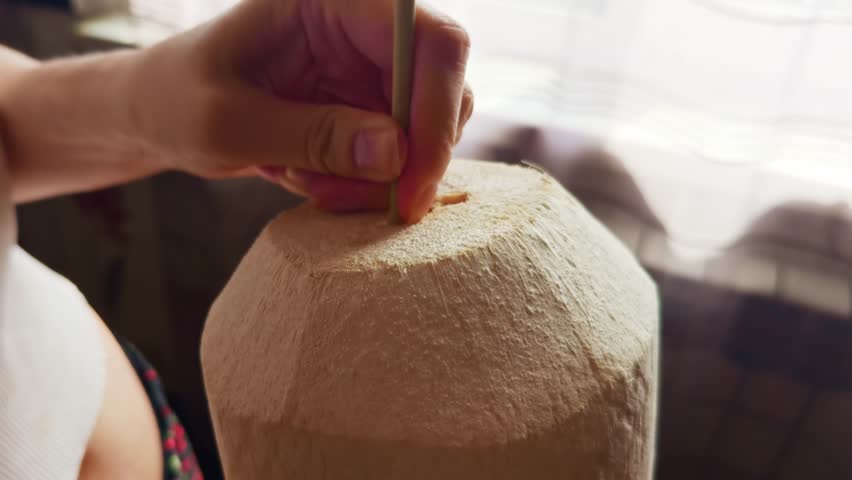 Coconut water. Hand inserting a straw into a fresh coconut for drinking coconut water. Natural refreshment scene indoors with soft daylight and healthy tropical hydration. Drinking coconut water. 