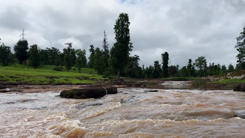 A river stream with green field behind it under cloudy sky