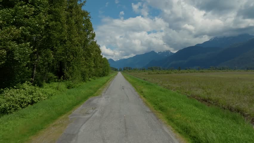A scenic drone view of a roadway winding through lush greenery, leading toward a distant mountain range under an open sky