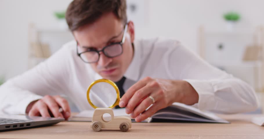 Focused young man in eyeglasses sitting at desk in office, examining wooden car with magnifier. Male car insurance expert writing data to notepad, searching and investigating on workplace. 4k video.