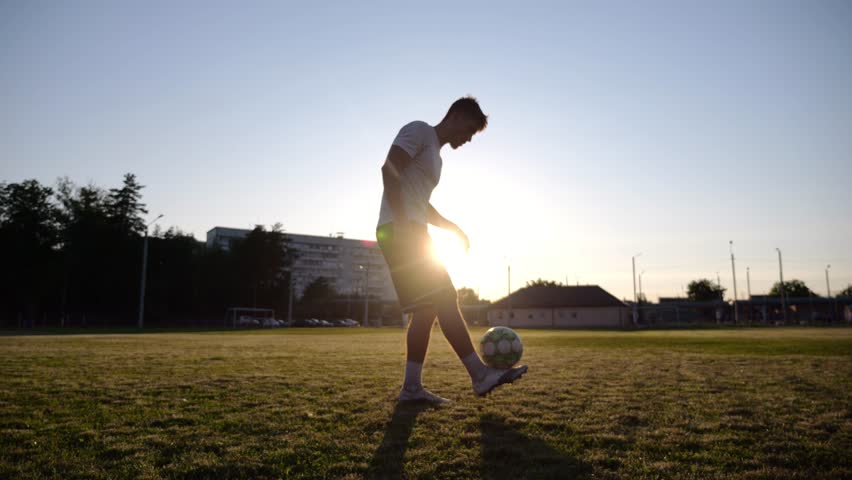 Professional footballer juggling soccer ball on stadium at sunset. Young man kicking ball at green field. Sportsman practicing tricks at meadow with sunlight at background. Freestyle football concept - Powered by Shutterstock - Get 15% off with code: PIKWIZARD15