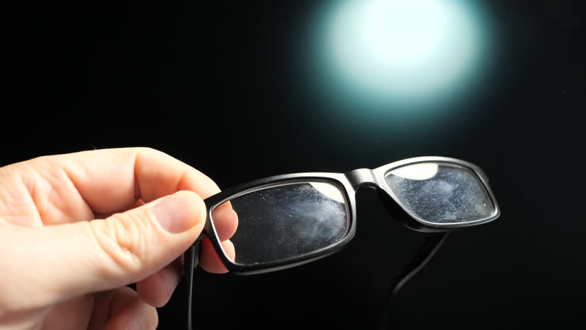 A closeup footage of a person cleaning the lenses of an eyeglasses with a cotton cloth, against a blurred bright light in the dark background