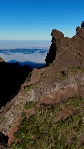 An aerial view of people ascending the hiking trail on Pico do Arieiro Mountain over clouds, with blue sky in the background in Madeira, Portugal
