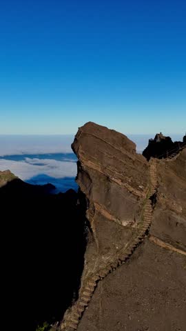 A drone vertical view of Pico do Arieiro Mountain with hiking trail over clouds, with blue sky in the background in Madeira, Portugal
