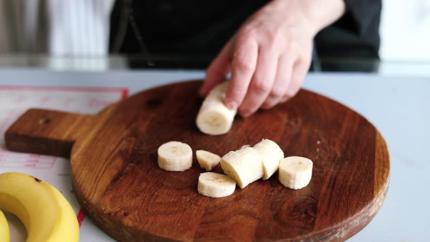 closeup of chopping fresh banana with knife on wooden cutting board.