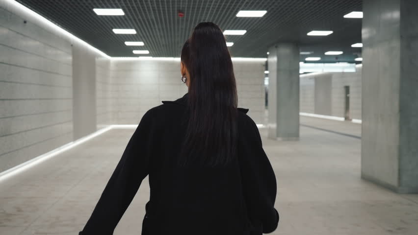 Following shot portrait of happy Latina female walking through modern station tunnel hall. Brunette woman wearing long black coat walks looking at camera and smiling. Futuristic architecture