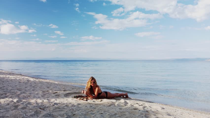 A blonde woman in a black bikini lies on a sandy beach, enjoying the sun and the view of the calm sea.