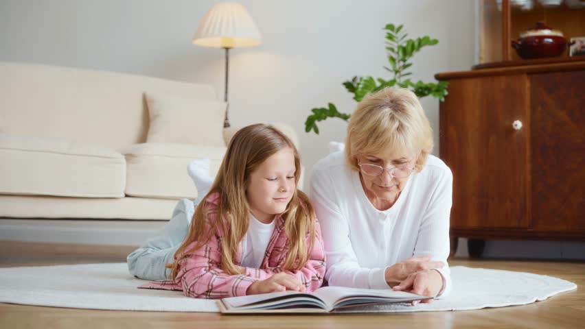 A warm scene of a 60-year-old grandmother and her 10-year-old granddaughter lying on the carpet, reading a book together. Bright cozy room, bonding through shared stories. High quality 4k footage