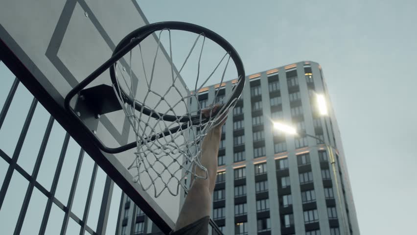 A young man is hanging on a basketball hoop and throwing a ball into it on an outdoor basketball court in the city. slam dunk