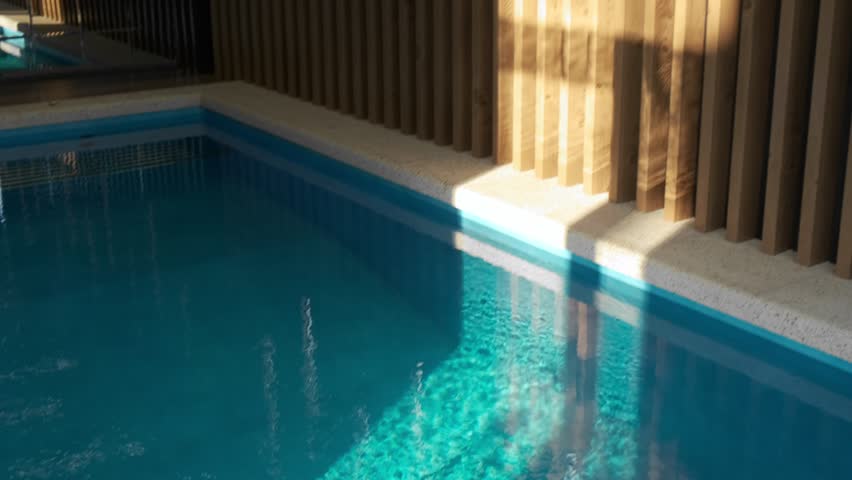 Decorative wooden wall near the edge of an indoor overflow pool. Modern lighting and water reflections create a cozy and relaxing atmosphere in a spa area
