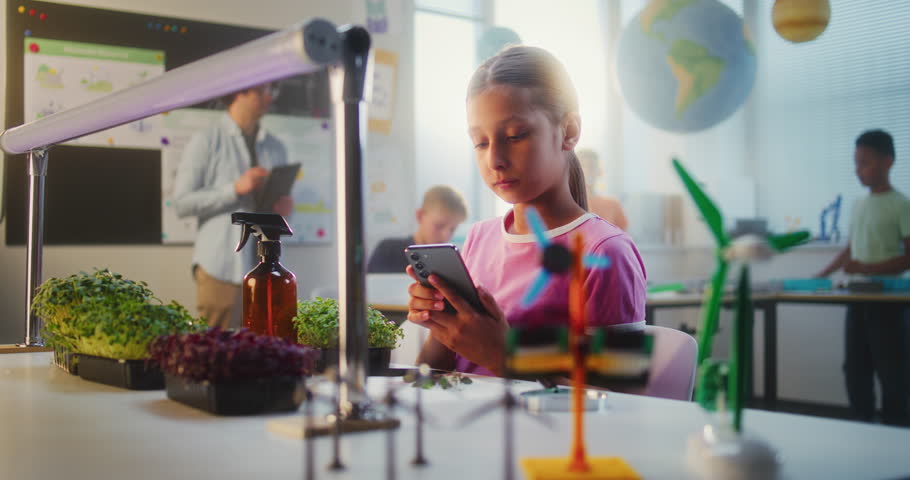 Talented Girl Sitting at Table and Using Smartphone, Conducting Biology Experiment on Plants with Tweezer and Magnifying Glass. Primary School Children During Lesson in Classroom. STEM Education.