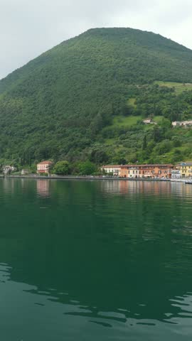 Serene sweeping drone view of a small boat approaching a charming lakeside village on Lake Isola in Northern Italy, with historic buildings and dramatic mountain backdrop.