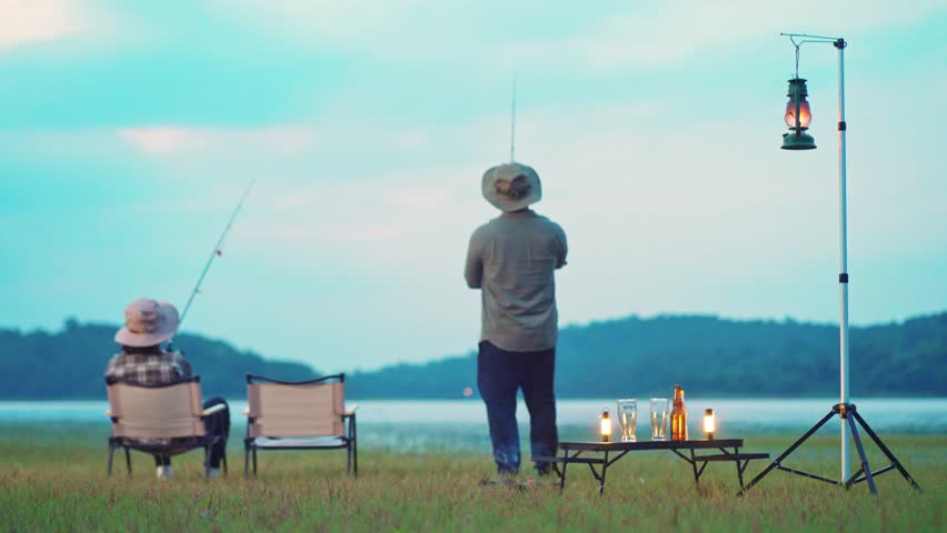 Romantic couple fishing together by the lake at dusk, sitting on camping chairs with lanterns and drinks nearby. Quiet outdoor lifestyle surrounded by nature. Selective focus only.