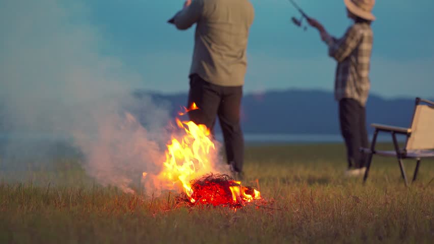 Romantic couple fishing together by the lake at dusk, sitting on camping chairs with lanterns and drinks nearby. Quiet outdoor lifestyle surrounded by nature. Selective focus only.