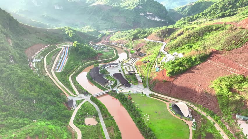 Aerial view of a scenic mountain valley in Moc Chau, Vietnam, featuring winding roads, terraced hills, and a river flowing through lush green landscapes. Captured in the early morning light