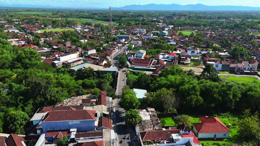 Medium-altitude aerial static shot focuses on a bridge spanning a green area within a densely populated suburban setting with red-tiled roofs and lush trees