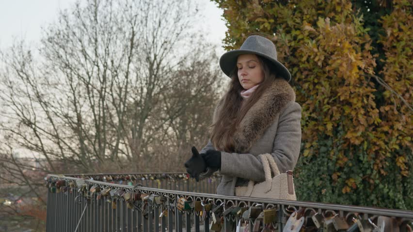 A pensive woman in a coat and hat stands on a bridge with love locks on an overcast autumn day. She looks out thoughtfully, with bare trees and foliage in the background.