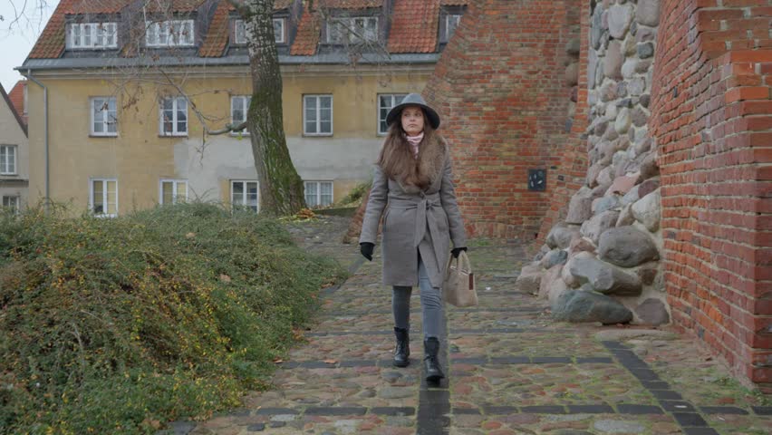A thoughtful woman walks on a wet cobblestone path along a historic wall. An atmospheric city scene after the rain on a cold, overcast autumn day in Warsaw.