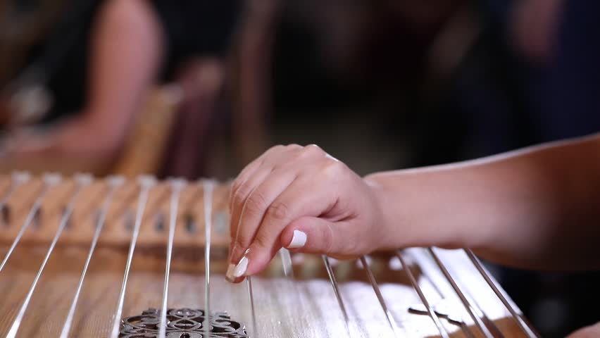 close-up shows a woman's hands playing an ornate wooden zither with plectrums. A microphone captures the beautiful sound of this traditional Middle Eastern folk instrument.
