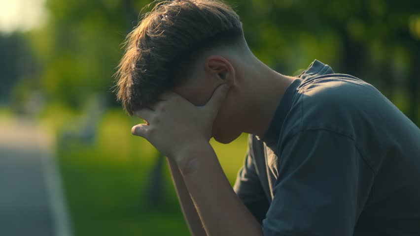 A young teenage boy sits quietly on a wooden bench, burying his face in his hands. The image captures a moment of deep thought, worry, and emotional weight.