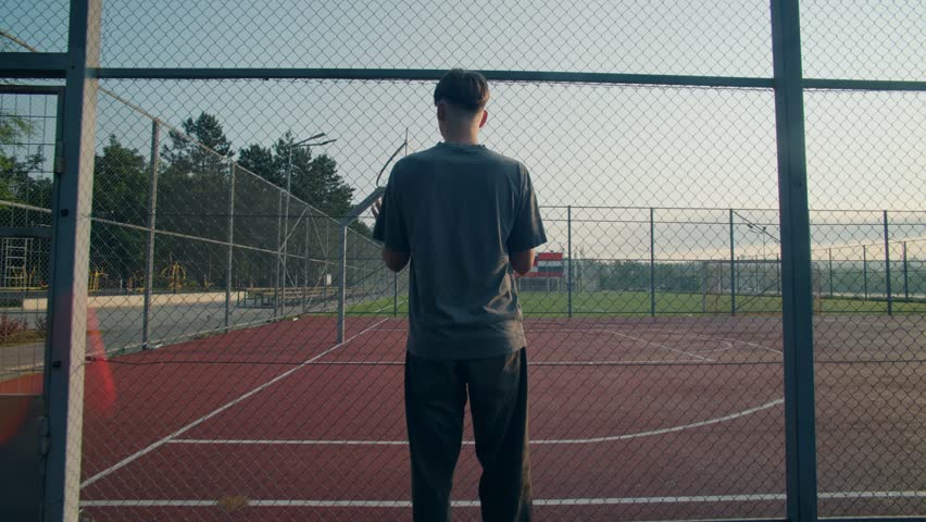 A teen stands outside a fenced basketball court at sunrise. The court is empty, but the vibe is full quiet, moody, and reflective of teenage emotion.