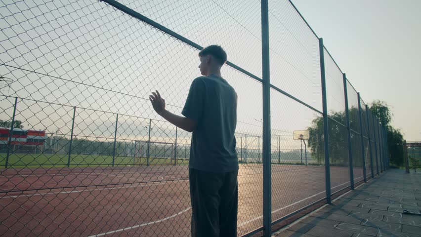 A teen boy gazes through a fence at an empty school basketball court. Caught in silence, he reflects a story of youth, isolation, and thought.