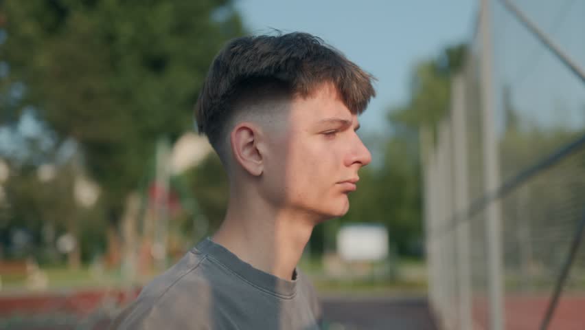 A teenage boy stands beside a chain-link fence, staring forward in silence with a pensive look. A sunny day frames a moment of deep thought, solitude, and reflection.