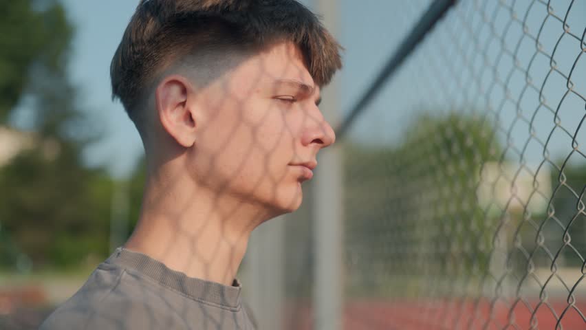 A teenage boy stands beside a chain-link fence, staring forward in silence with a pensive look. A sunny day frames a moment of deep thought, solitude, and reflection.