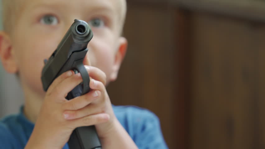 The hands and face of a young boy are partially obscured by a black toy gun that he is holding with both hands, with the barrel pointed towards the viewer, indoors.