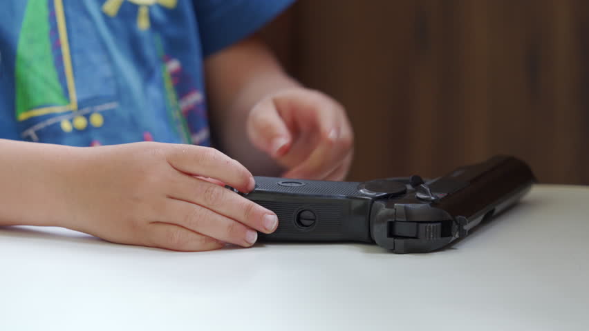 The hands of a young child are seen holding and manipulating a black toy gun, which rests on a white table indoors.