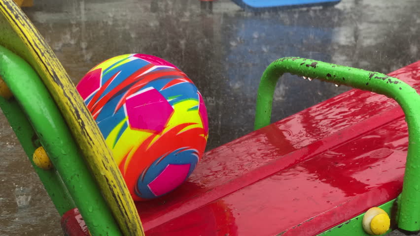 A vibrant ball rests on a wet teeter-totter, glistening under a gentle rainfall. The scene captures a moment of playful abandonment in what appears to be an outdoor playground during a summer shower.