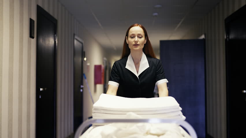 Young hotel maid pushing trolley with clean linen for servicing guest rooms