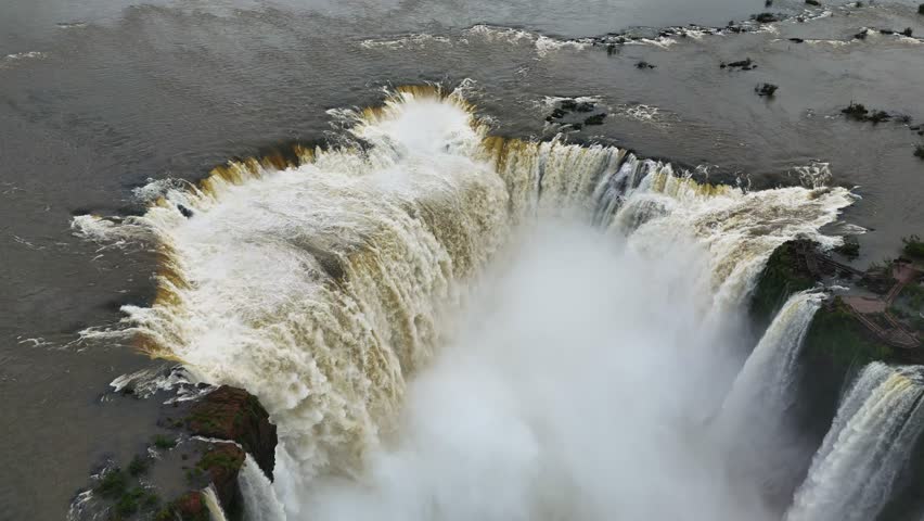 Aerial view moves above thunderous Iguazu Falls in Brazil Powerful cascades drop over rocks, with mist rising and green cliffs visible The scene is dramatic, wild, and filled with natural energy
