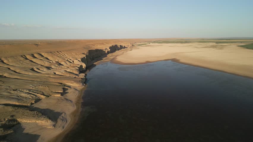 Eroded cliffs overlook the meandering Amu Darya river. Broad sandy floodplains stretch far into the horizon. Vegetation patches line the water's edge, enhancing the desert's contrast