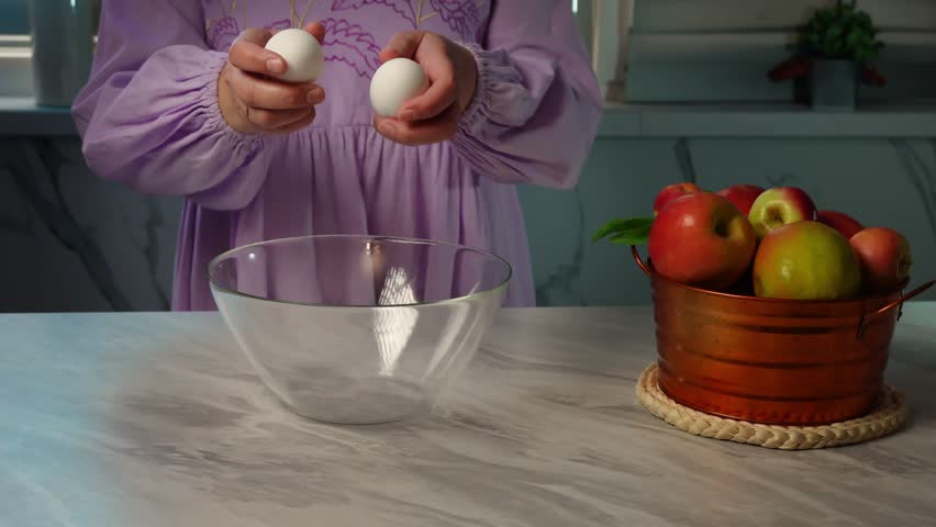 Woman in purple dress cracks egg into glass bowl on marble countertop