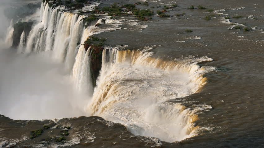 Aerial view shows Devil's Throat as torrents of water plunge over a massive cliff. Mist fills the air in Brazil as currents converge. Lush vegetation clings to rock faces creating a unique landscape
