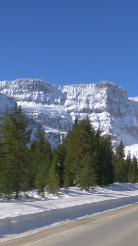 POV: Driving along the famous Icefields Parkway route on a sunny winter day. Evergreen forest covers the rugged landscape under the snowy Canadian Rocky Mountains. Scenic route across Alberta, Canada.