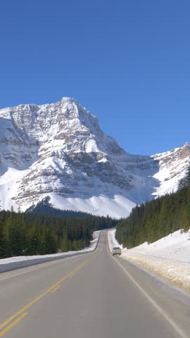 POV: Driving along the famous Icefields Parkway route on a sunny winter day. Evergreen forest covers the rugged landscape under the snowy Canadian Rocky Mountains. Scenic route across Alberta, Canada.