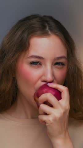 Portrait of Young Woman Eating Healthy Food and Looking at Camera. Real Person on Diet Consuming Fresh Red Apple as Lunch Snack. Playful Female Human Inside Cozy Living Room Alone with Organic Meal