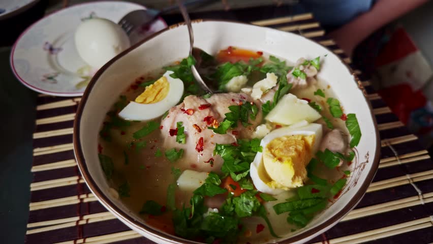 Bowl of homemade chicken soup with boiled egg, vegetables, and fresh herbs. A spoon rests inside the bowl. Hearty and healthy meal served on a bamboo mat, with a boiled egg. Dieting chicken soup bowl 