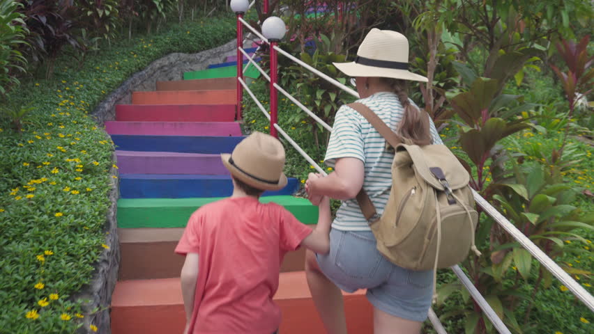 Female tourist and her son are going up the steps. Colorful stair. Woman and little boy in hats at a scenic garden. Traveling with child. Family vacation. Trip with backpack.