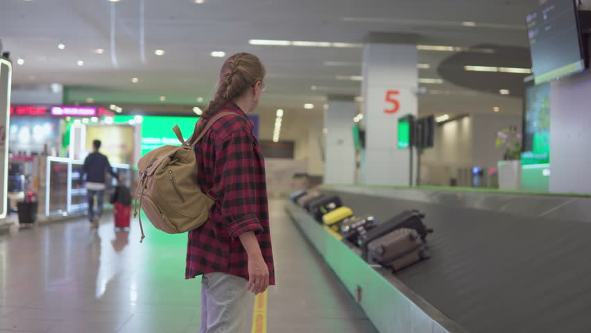 Female tourist with backpack is waiting her suitcase near luggage conveyor belt at arrival area of passenger terminal of airport. View of suitcases on baggage carousel. Solo traveler. Single vacation.