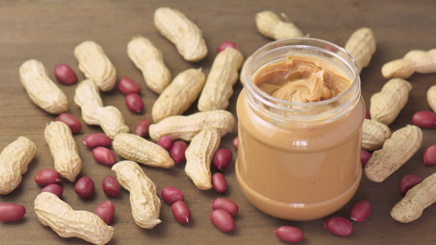 Female hands taking a spoon of tasty peanut butter from a jar. Macro shot. Groundnut on wooden table. Sweet organic food.