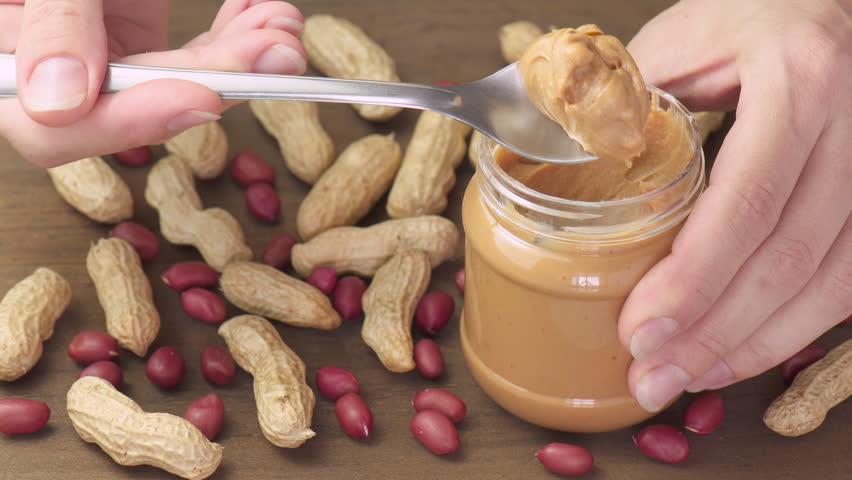 Female hands taking a spoon of tasty peanut butter from a jar. Macro shot. Groundnut on wooden table. Sweet organic food.